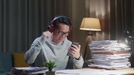 hombre asiático con auriculares escuchando música en su teléfono inteligente después de trabajar con documentos en casa