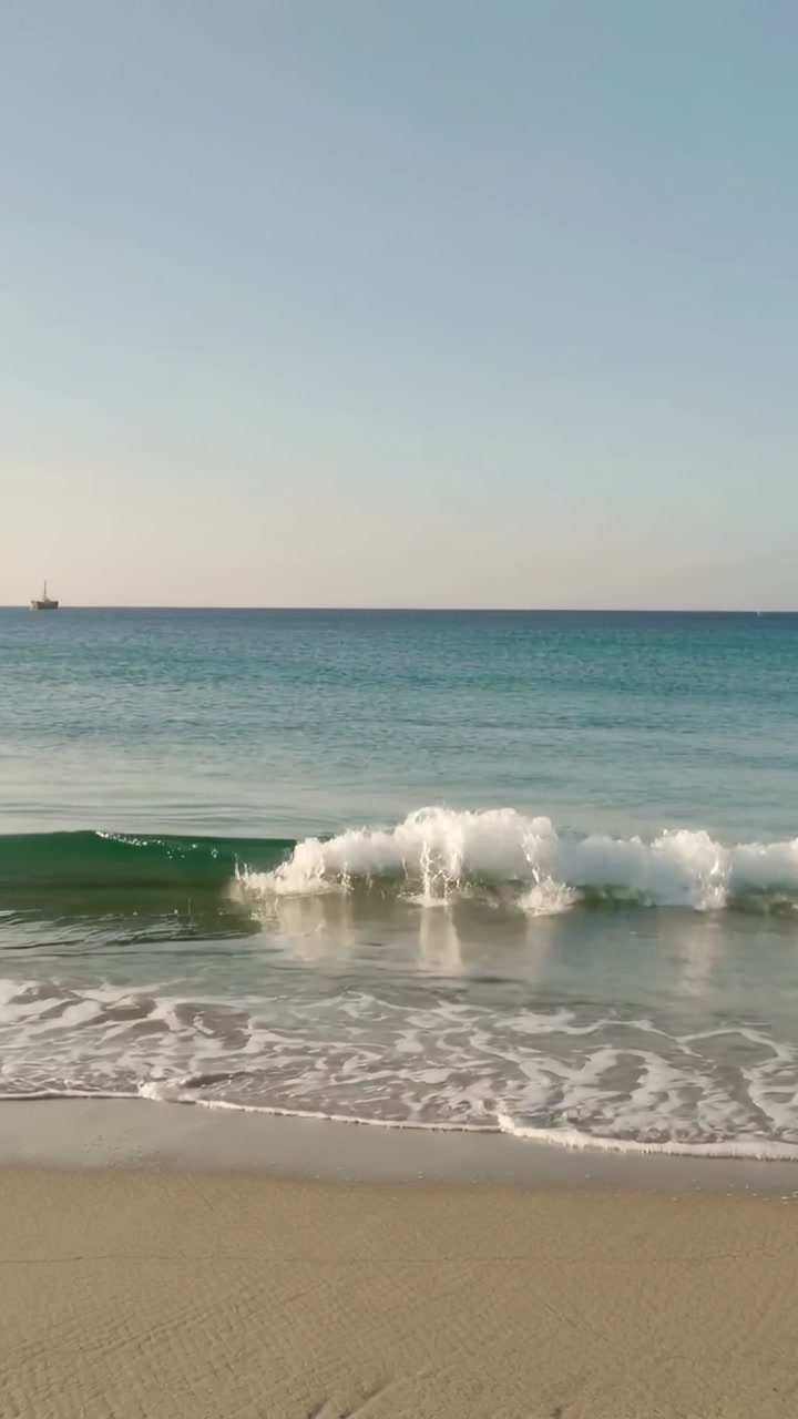 Vertical video: Incoming swells pushing waves toward wet sand, forming foam near left horizon boat