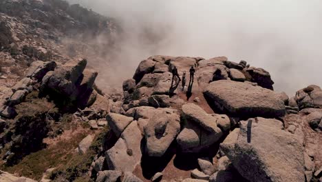 Aerial-tilt-shot-of-men-at-rocky-mountain-peak-with-clouds-in-background