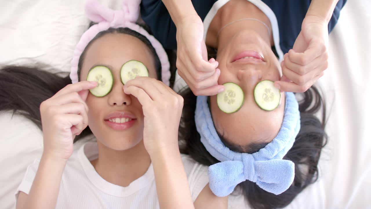Biracial teenage girl pals lie down at home, holding cucumber slices on eyes