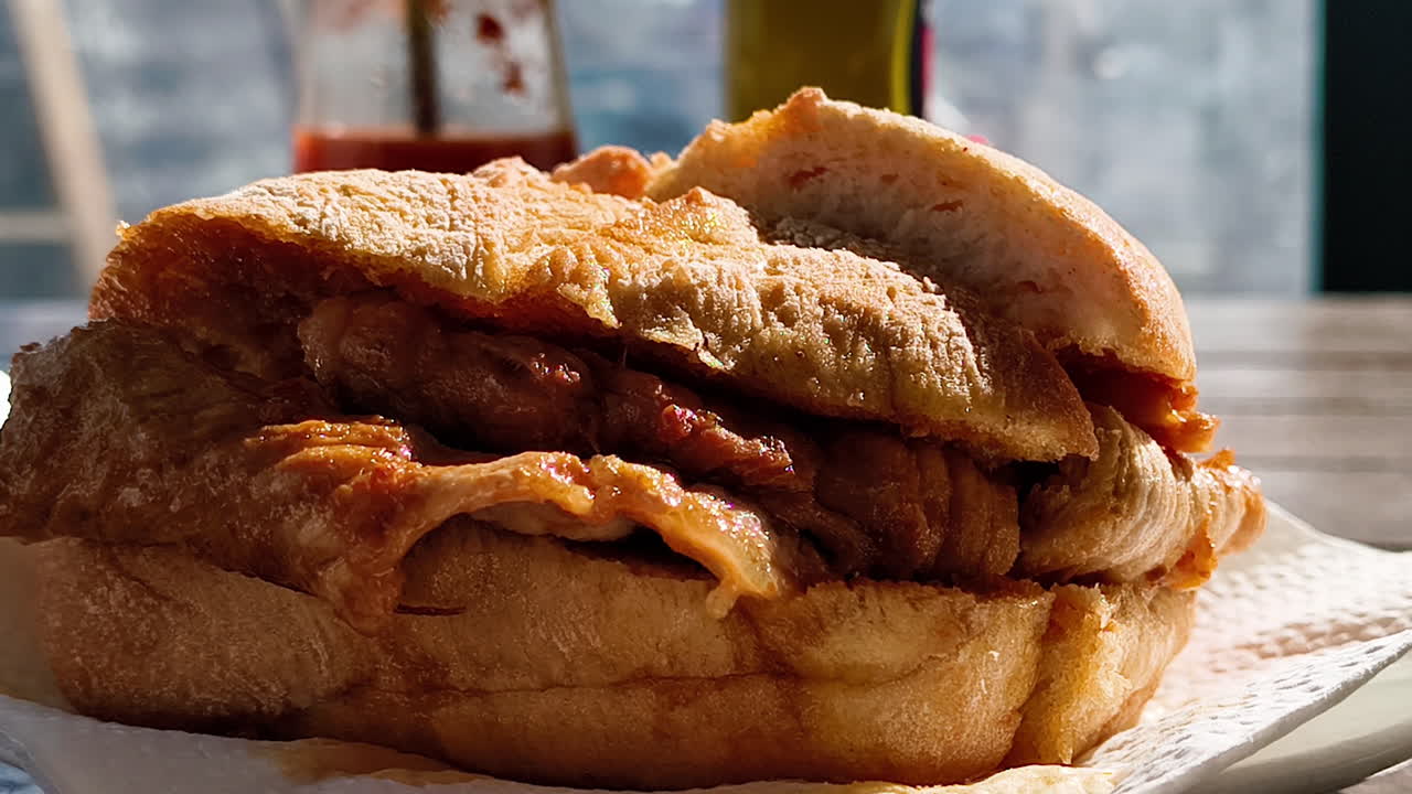 A close-up of a meat sandwich on a white paper napkin, with a bottle of spicy and mustard in the blurry background. The sandwich is filled with thick, juicy meat and has a golden-brown crust.