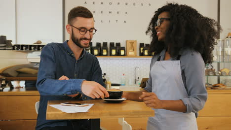 young caucasian man sitting at the table and using smartphone in coffee shop, then african american waitress brings him a cup of coffee