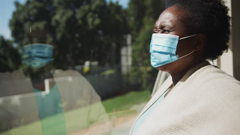 Senior-african-american-woman-wearing-face-mask-standing-by-window