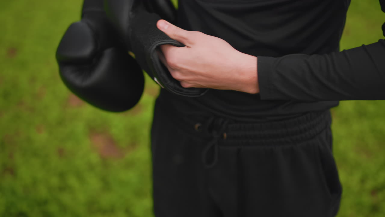 Boxer wickelt Bandagen vor dem Training auf einem grasbewachsenen Trainingsplatz, schwarze Handschuhe mit Klettverschluss, Nahaufnahme von Händen und Handgelenk, ruhige, fokussierte Energie und einsame Vorbereitung auf das Training