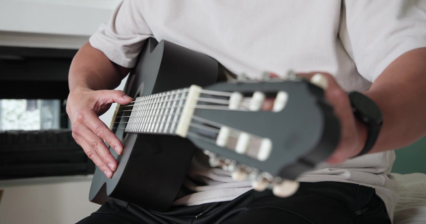 Senior Chinese male plucking strings checking tuning adjusting pegs and playing black guitar on bed