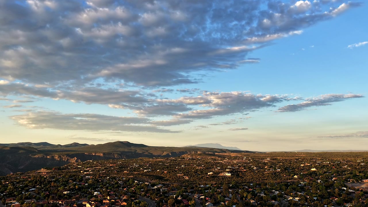4K UHD drone-timelapse som fanger mykt daggrylys over en forstadsby i en kløft. Raskt bevegelige, teksturerte skyer passerer over en blå himmel over det vidstrakte nabolaget, platået og fjerne fjell