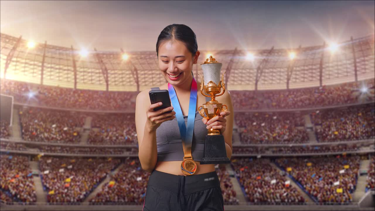 mujer asiática con una medalla de oro sosteniendo un trofeo de oro y usando un teléfono inteligente en el estadio