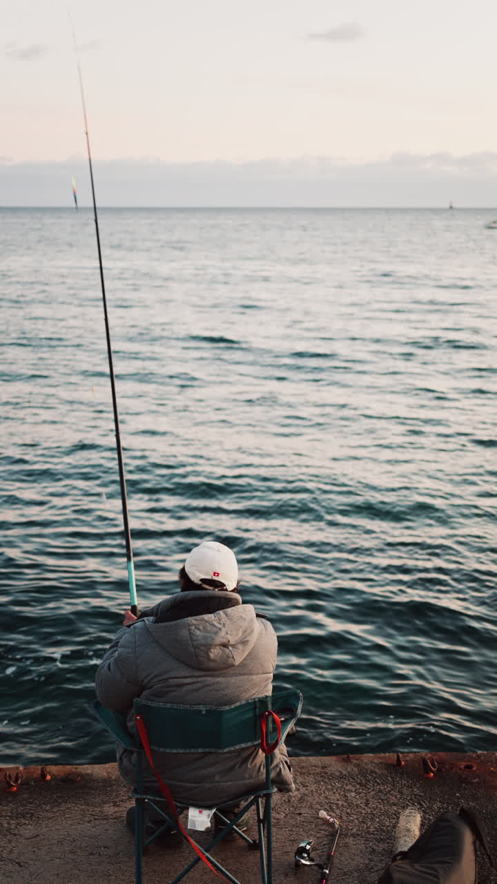 Man in winter clothes on a chair fishing in the sea in the south of France. Vertical