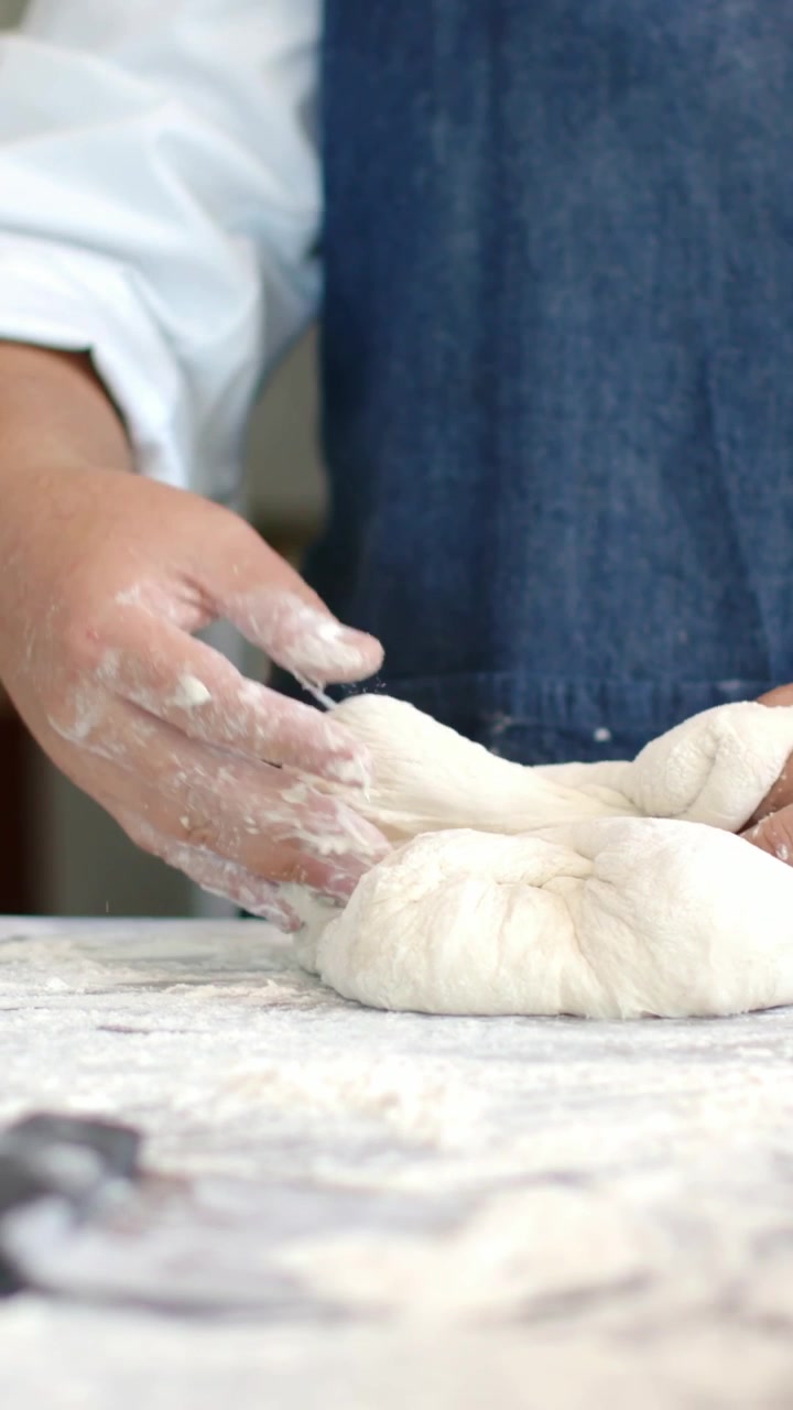 Vertical video: Woman in denim apron lifting dough, shaping dough into round to bake on floured top