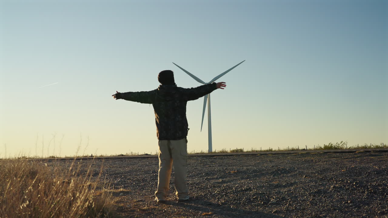 A person stands with arms open facing a windmill on the horizon beneath a clear blue sky