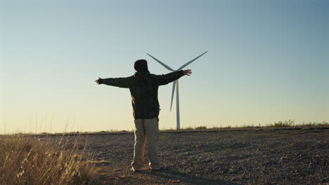 a person stands with arms open facing a windmill on the horizon beneath a clear blue sky