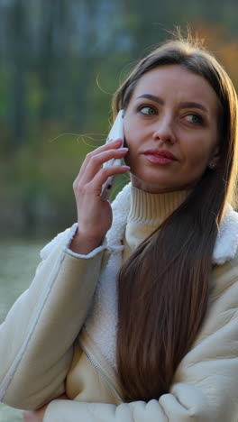 pleasant-looking caucasian woman waiting for a response on her phone. lady stands on the bridge over the river in autumn. vertical video
