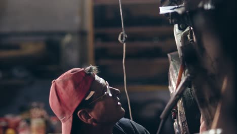 Male-worker-with-glasses-and-red-cap-on-at-mechanical-hangar-adjusts-something-on-metal-construction-with-a-hammer.