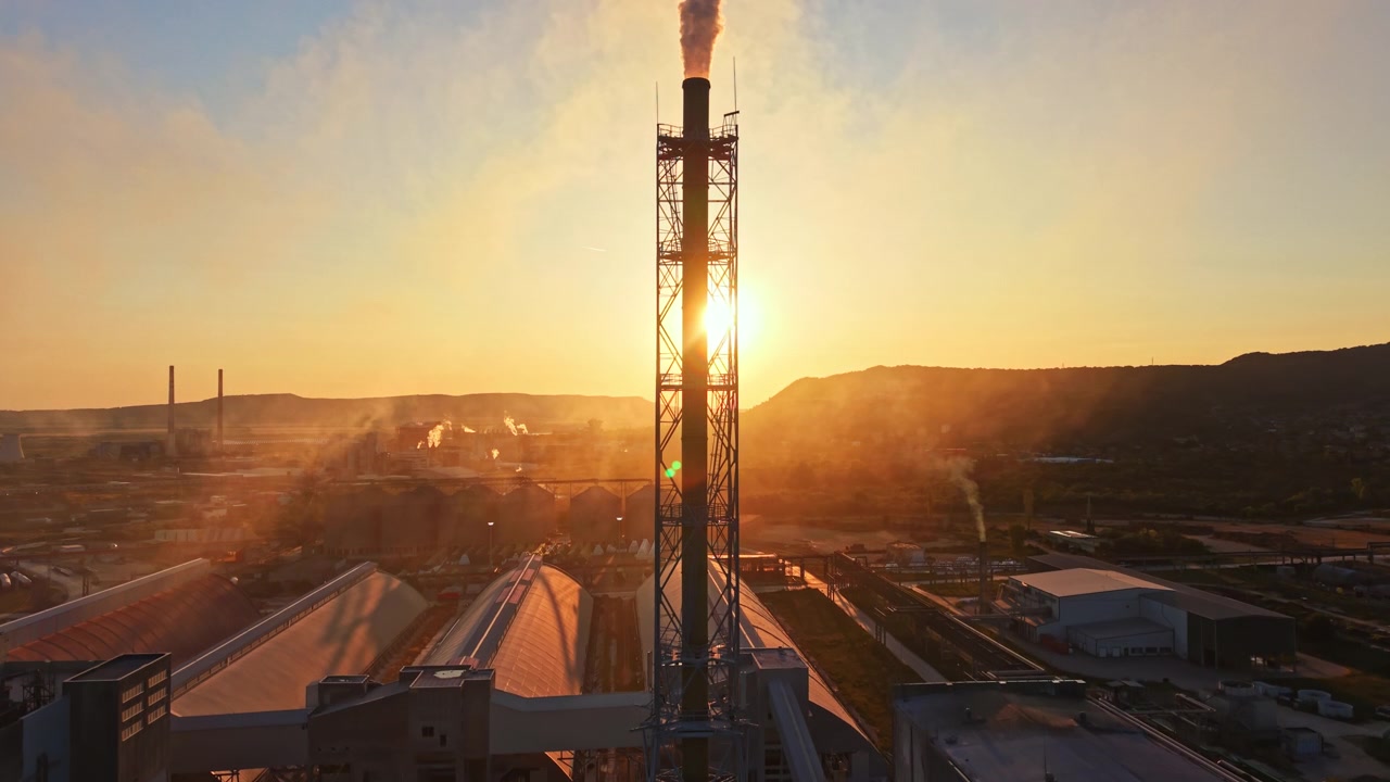 Vista al tramonto di una zona industriale con un'alta torre e fumo che sale sullo sfondo.