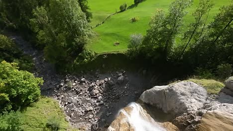 Fast-pace-aerial-descend-over-rugged-mountain-waterfall-to-green-valley-meadow