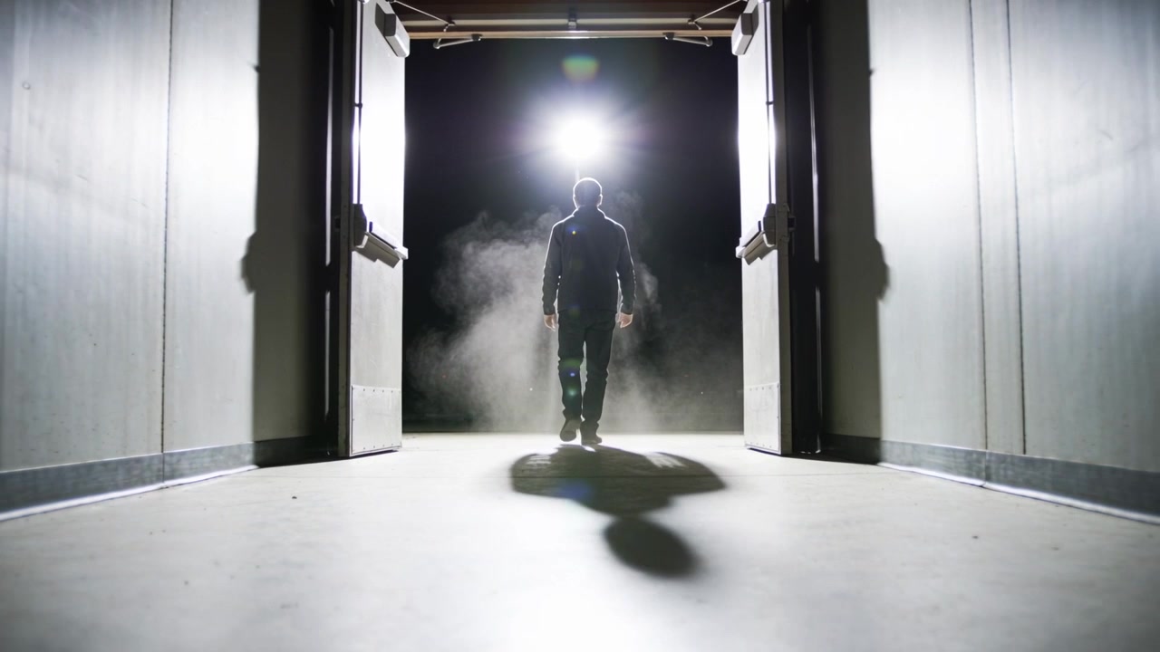 Lopend man in donker jack stapt door open dubbele deuren een gang in, met tegenlicht en mist.