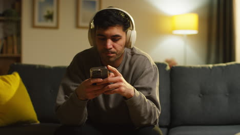 young man spending evening at home sitting on sofa wearing wireless headphones streaming music from mobile phone answering call 1