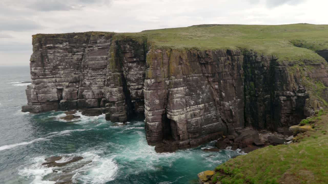 Waves gently crash over rocks and against the base of a tall sea cliff in the deep teal coloured ocean while seabirds fly around the cliffs of a colony of guillemots on Handa Island, Scotland