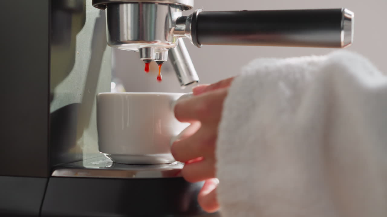 el café preparado gotea desde la máquina hasta la taza en primer plano. una mujer en bata de baño espera llenando la taza con café caliente de la cafetera en la cocina. concepto de ritual matutino