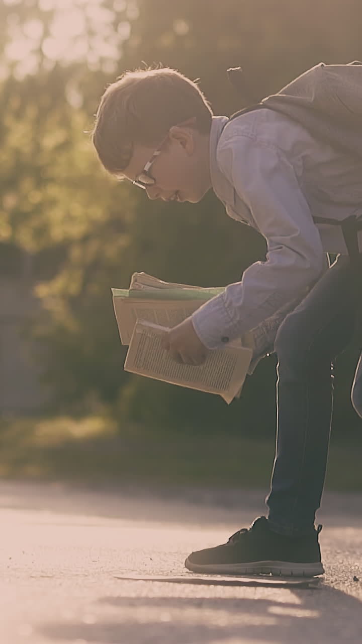 young guy with black schoolbag and in glasses gathers books and sheets on asphalt road against green trees slow motion