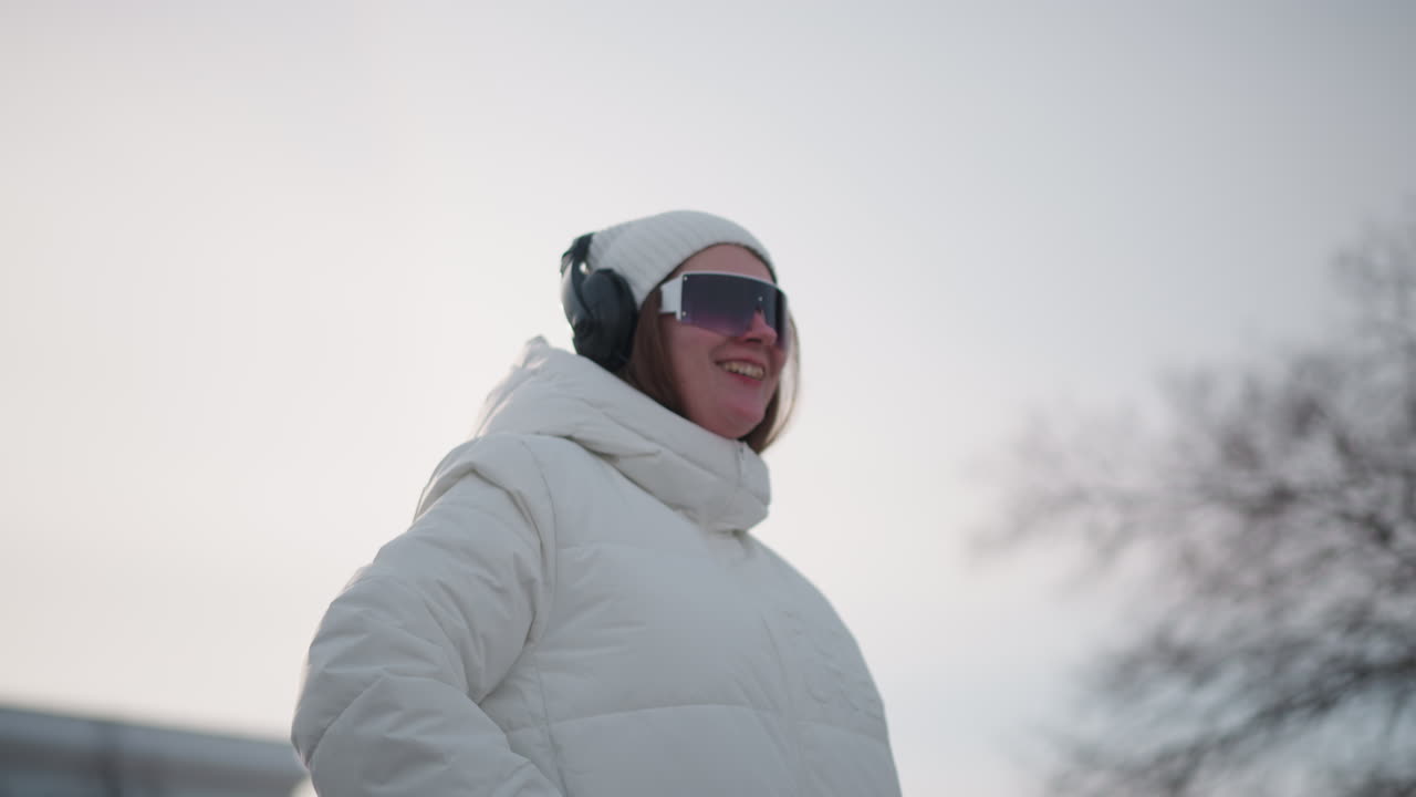 Student dancing in snow smiling joyfully while wearing puffer jacket knit beanie and headphones arms outstretched captured outdoors against white winter landscape conveying playful motion energy