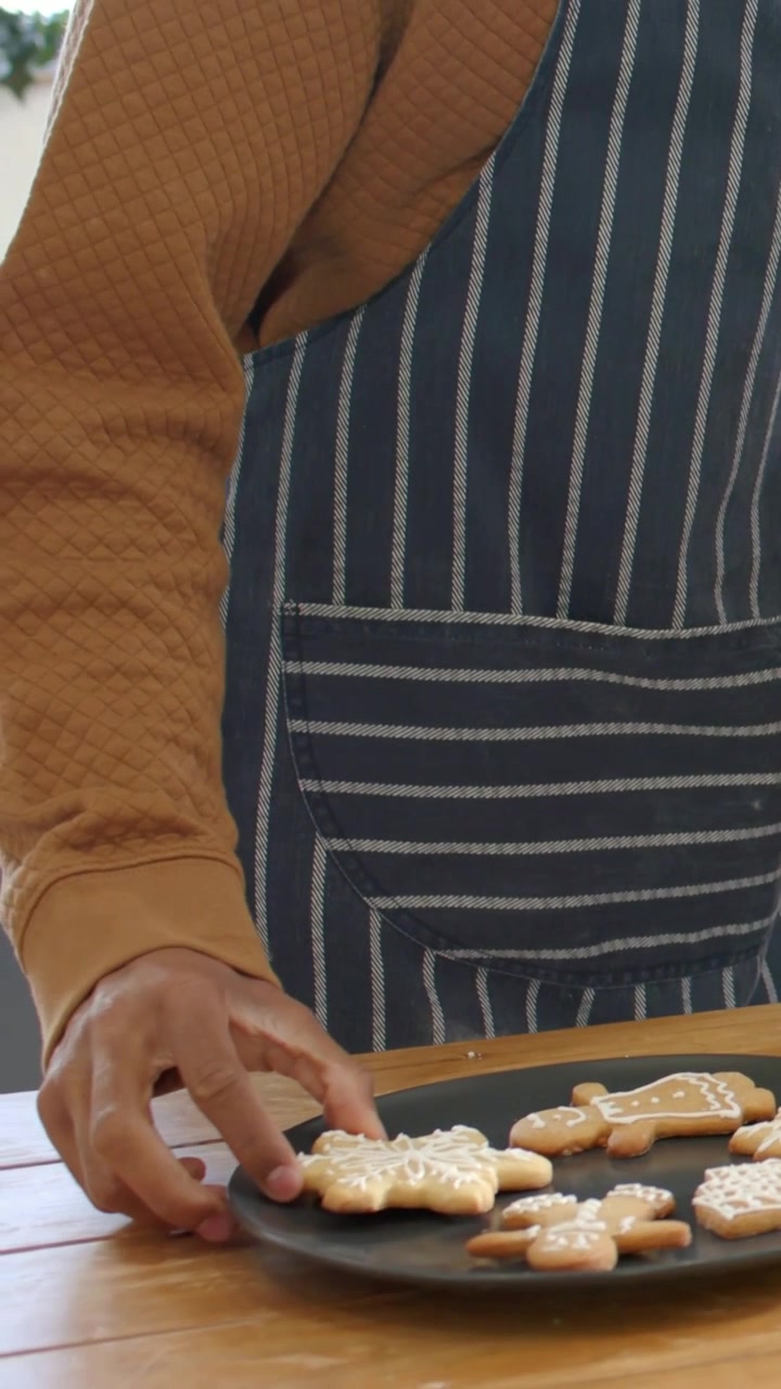Vertical video: Hand reaching, apron-clad mustard-top woman nudging cookies on black plate to serve