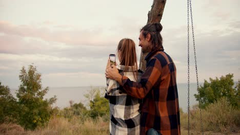 Shooting-from-behind:-A-guy-in-a-checkered-shirt-hugs-and-caresses-his-girlfriend-in-a-checkered-shirt-while-she-takes-a-picture-of-a-sea-landscape-on-her-phone.-Rest-in-the-country-house