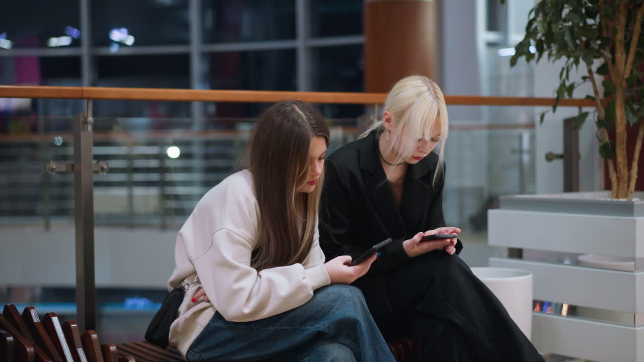 Two sisters walking indoors holding phone with tired expression in modern mall environment showing casual lifestyle fashion candid mood urban atmosphere