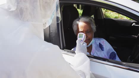 trabajador de la salud con ropa de protección midiendo la temperatura de una mujer mayor afroamericana en un coche