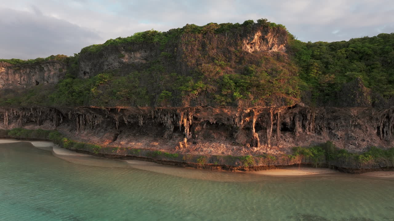 Drone close-up of limestone cliffs in Lekini Bay, Ouvéa, New Caledonia, bathed in golden sunset tones over clear waters