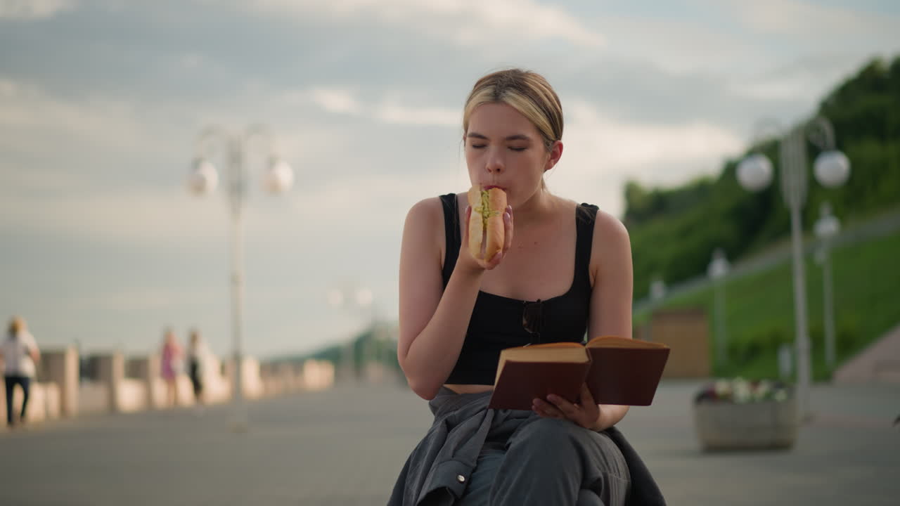 소시지 롤을 먹고 읽는 동안 한 손에 책을 들고 다리를 은 채 야외에 앉아있는 여성, 배경에는 램프 기둥과 멀리서 는 사람들이 있습니다.