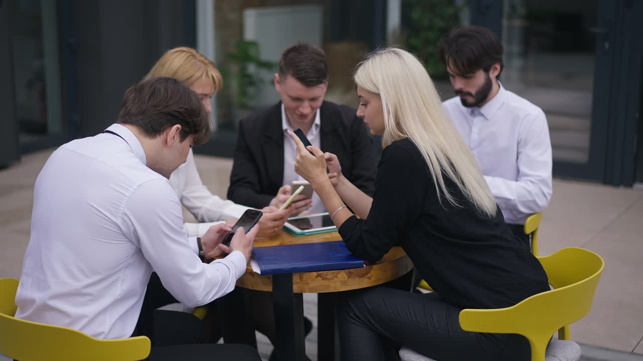 Group absorbed colleagues sitting at round table on office terrace surfing Internet on smartphones. Caucasian men and women analyzing e-market planning strategy outdoors. Slow motion.
