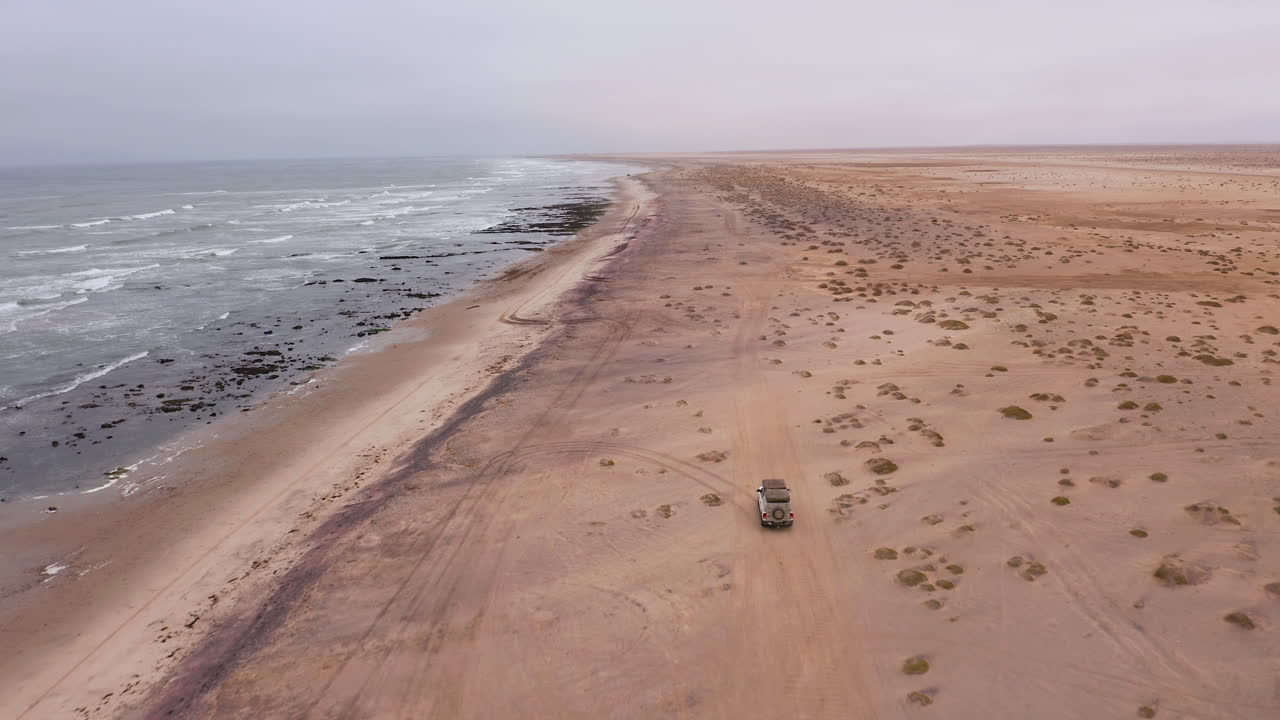 Aerial: Following of an off-road vehicle cruising along the wide sandy beach near Swakopmund, Namibia, under a grey overcast sky