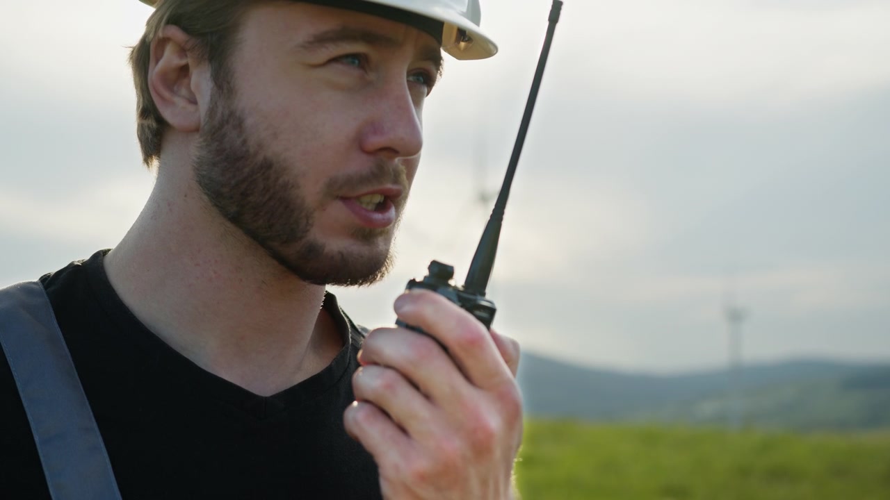 Uomo con cappello e camicia nera che tiene un walkie-talkie, con turbine eoliche sullo sfondo, simbolo di energia rinnovabile
