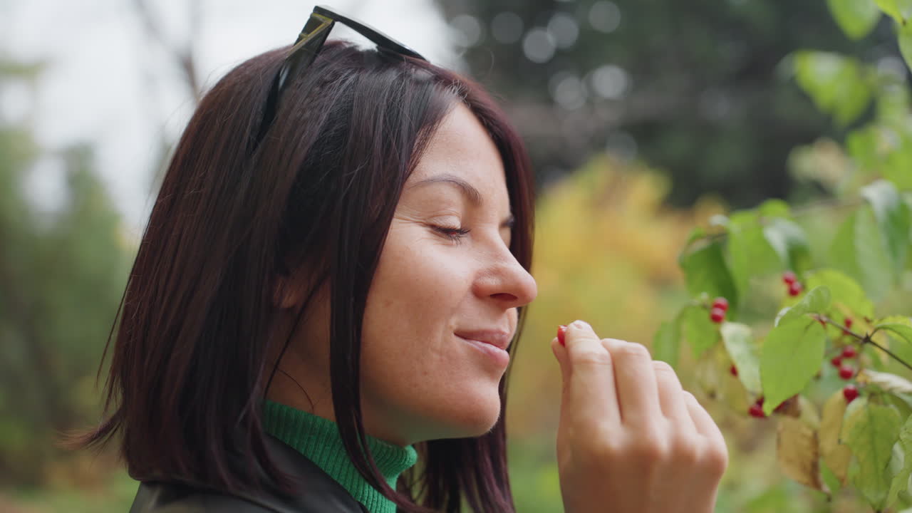 Donna che annusa bacche selvatiche in una foresta, giacca di pelle e occhiali da sole sulla testa, profilo ravvicinato con occhi chiusi e un sorriso accennato, fogliame autunnale e brezza leggera, atmosfera serena con texture terrosa e slow motion