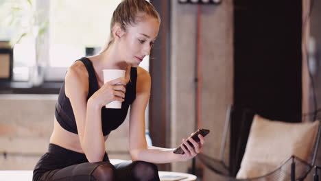 Female-Athlete-Using-Mobile-Phone-And-Having-Drink-While-Relaxing-After-Workout-In-Gym