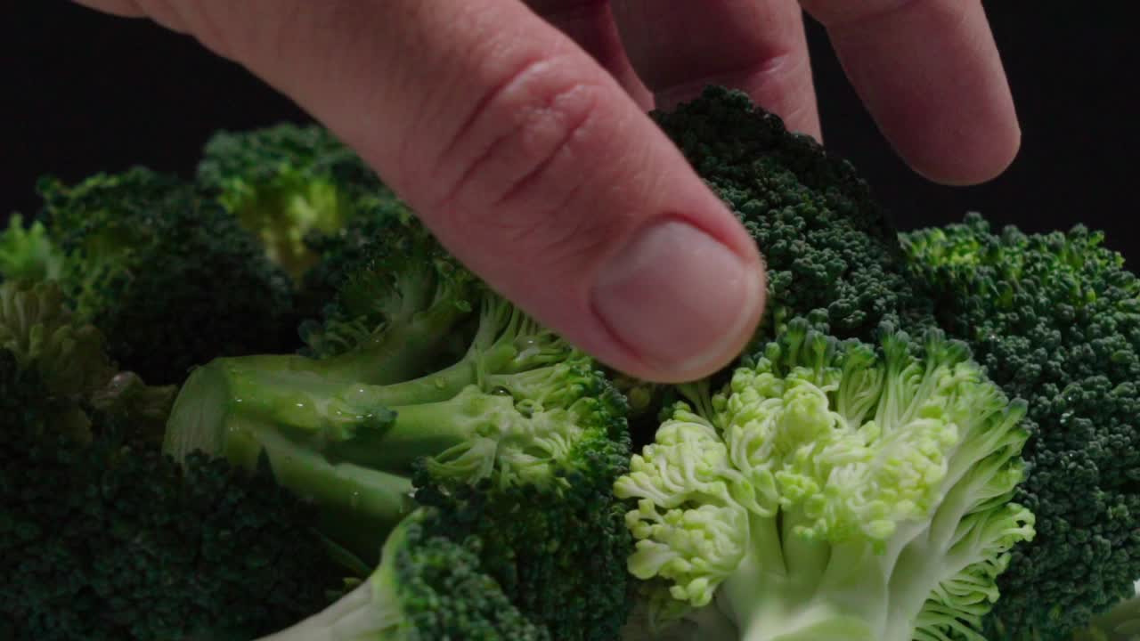 Hand Takes Broccoli Piece from Pile, Close Up on Black Background