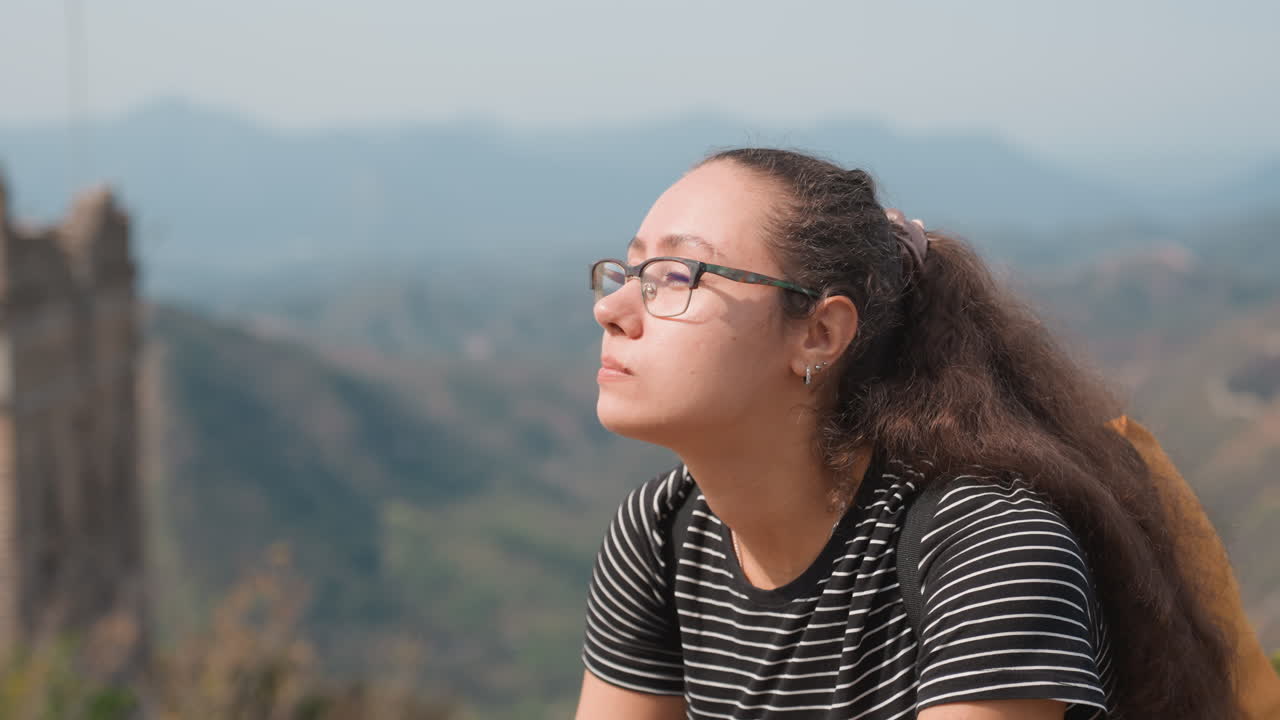 Ritratto ravvicinato di donna pensierosa che guarda verso creste distanti, rovine e un muro sinuoso sullo sfondo, luce naturale soffusa, atmosfera tranquilla e introspettiva, maglietta a righe casual e occhiali, brezza leggera