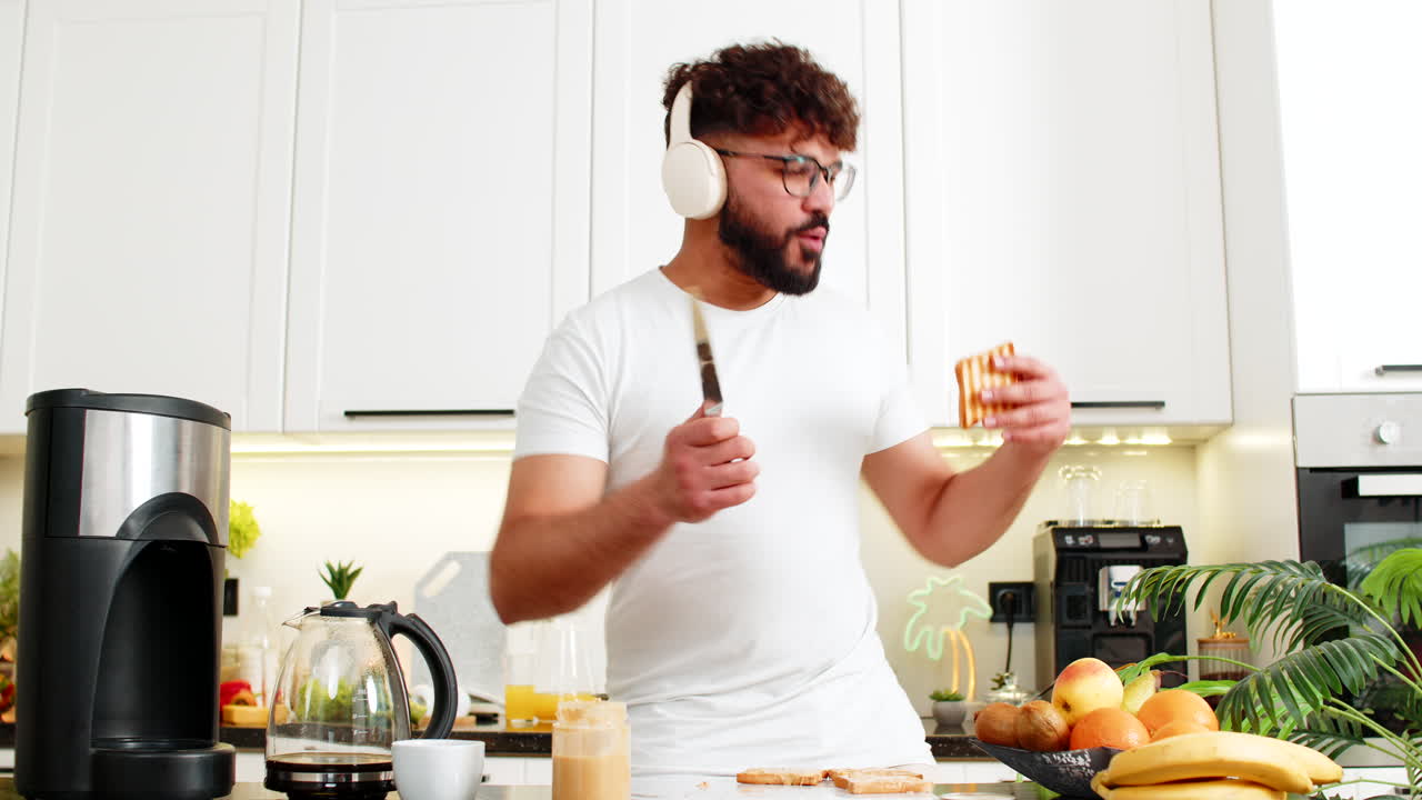 Uomo adulto arabo che balla con le cuffie mentre prepara toast con burro di arachidi per colazione nella cucina di casa