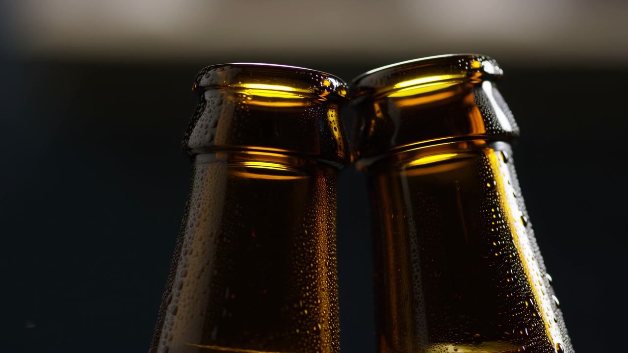 Primo piano ravvicinato da angolazione bassa che mostra due colli di bottiglia di vetro ambrato con i bordi che si toccano, coperti di condensa, su uno sfondo scuro sfocato con luce direzionale calda e riflessi sottili.