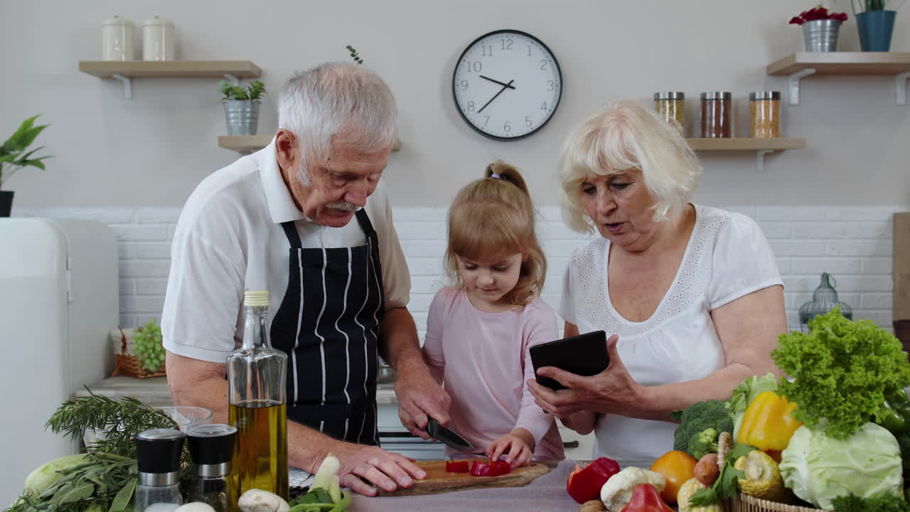 Senior grandparents couple with digital tablet and granddaughter cutting vegetables in kitchen
