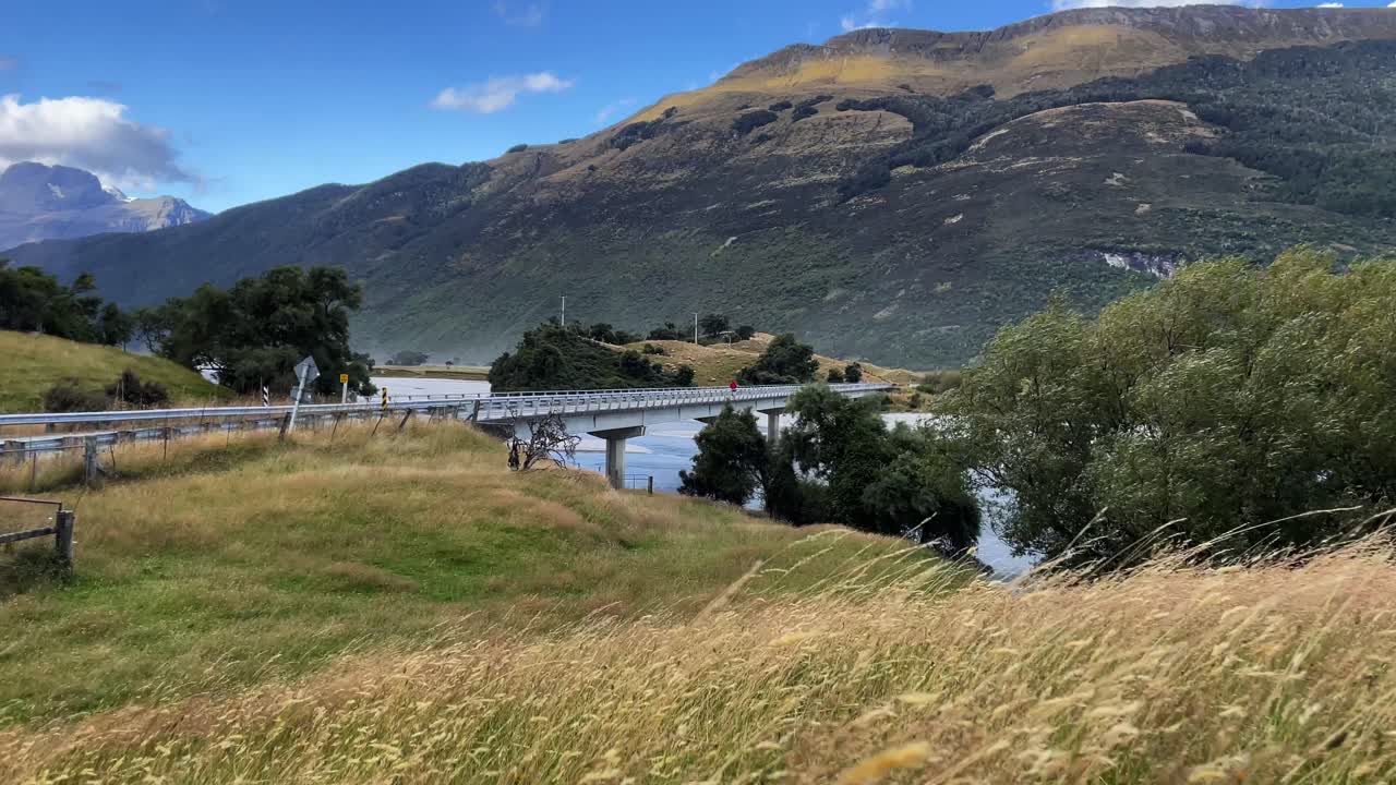hombre corre a través del puente en un día de viento, con montañas y cielo azul, nueva zelanda