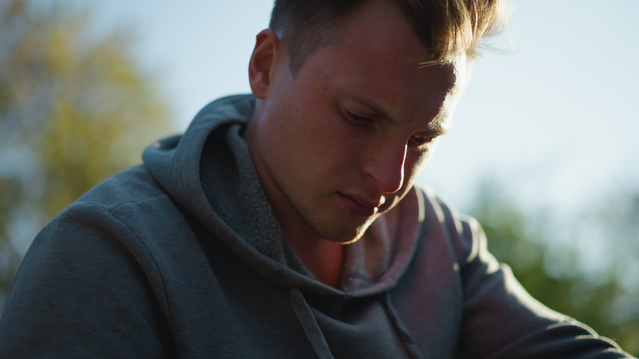 어두운 회색 후드 스웨트 셔츠를 입은 젊은 남자의 클로즈업, 집중적이고 사려 깊은 표정으로 내려다보고. 배경에는 녹색 잎자루가 흐릿합니다.