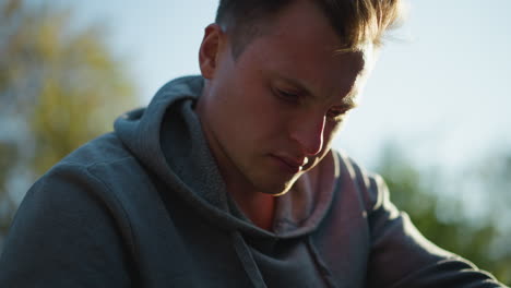 close-up of young man outdoors wearing dark gray hooded sweatshirt, looking down with concentrated, pensive expression. blurred green foliage in background