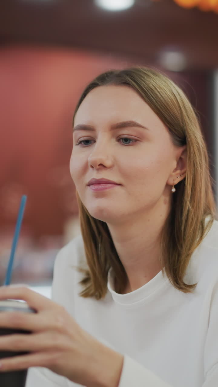 Lady in white shirt sips coffee from cup with subtle smile while seated in shopping mall cafe, background features warm lighting, greenery, and blurred restaurant signs