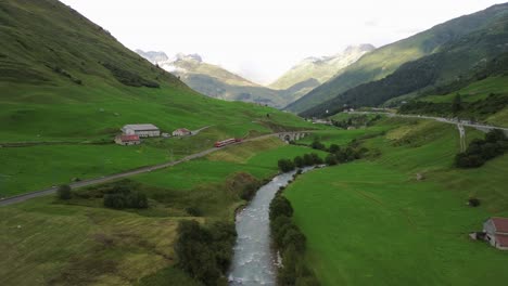 tren pasando por los pintorescos alpes suizos con pequeño puente y río al lado de las vías durante el verano en el paso de furka, filmado desde un dron en 4k desde una distancia