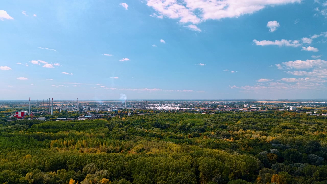 Industrial refinery beyond a broad expanse of woodland. An aerial panorama shows factory towers and a distant white plume above trees beneath a blue sky.