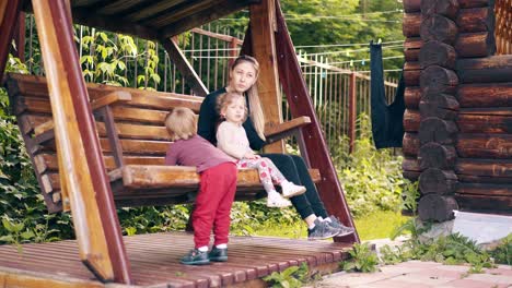 young pretty mother with children swinging on a wooden swing near the summer house on vacation