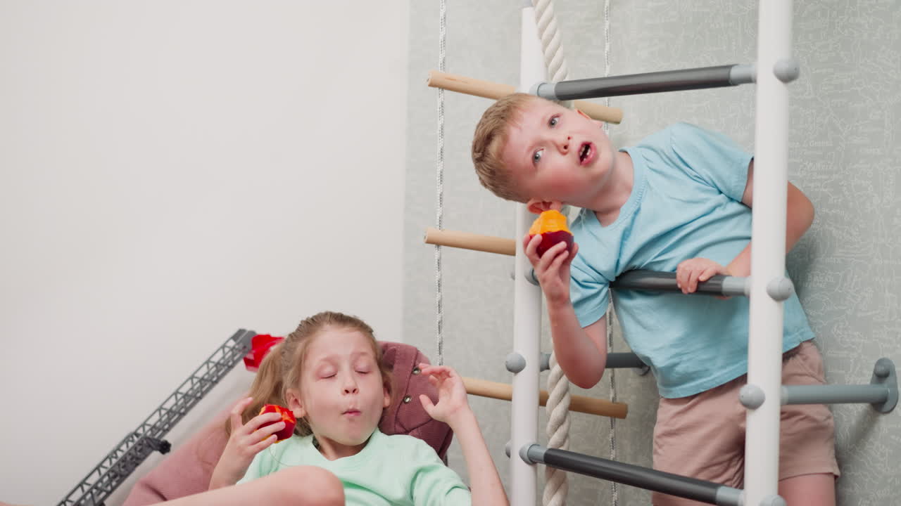 Children relaxing indoors enjoying juicy fresh fruits as boy hangs playfully eating vibrant mango from indoor ladder while girl lounges comfortably on bean bag chair savoring delicious fruit