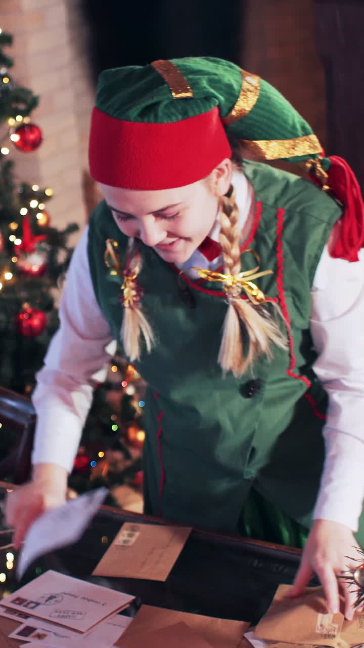 Elf girl in a green suit is looking for a letter on a table in a room with a Christmas tree and decorations. Vertical video